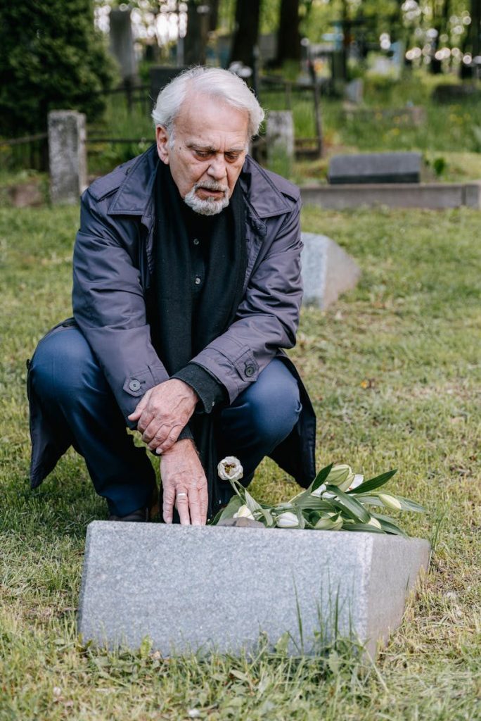An elderly man kneels in reflection at a gravestone, symbolizing loss and remembrance.